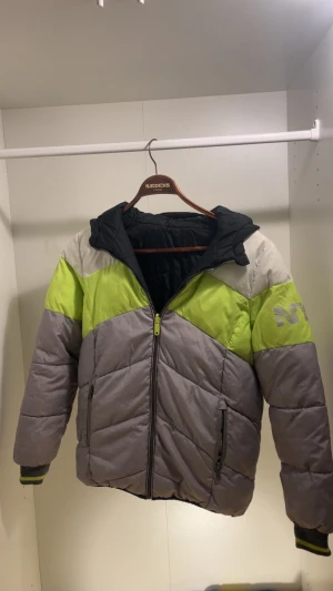 NYC pufferjacka i grått, lime och vitt - Fet pufferjacka från NYC med cool färgblock-design i grått, limegrönt, vitt och svart. Jackan har huva, dragkedja framtill och två fickor med dragkedja. Stort NYC-tryck på ena ärmen. Perfekt för kalla dagar när du vill sticka ut.