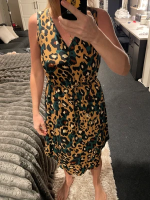 Leopardmönstrad ärmlös klänning - Snygg ärmlös klänning med leopardmönster i beige, svart och grönt. Klänningen har v-ringning, knyt i midjan och en avslappnad passform som ger en cool vibe. Perfekt för dig som gillar att sticka ut med djurmönster och vill ha något bekvämt men trendigt.