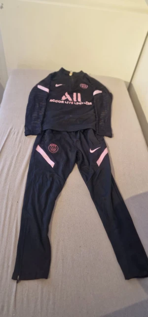 Svart PSG x Nike långärmad träningströja - Svart långärmad träningströja från Nike och PSG med rosa detaljer och loggor. Tröjan har half zip och är tillverkad i ett sportigt material som passar perfekt för fotbollsträning. Snygg design med rosa Nike-logga och PSG-märke på bröstet.