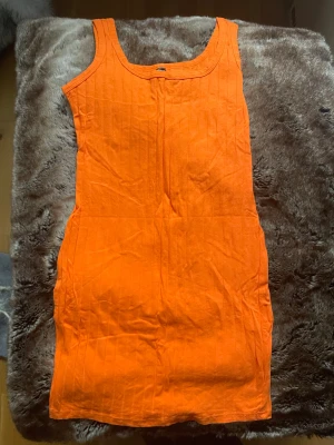 Orange  kortklänning - Säljer en snygg orange kortklänning med ärmlös design och rund hals. Klänningen har en enkel, rak passform och är tillverkad i ett lätt bomullsmaterial med subtila vertikala ränder. Perfekt för dig som gillar färgstarka och stilrena plagg.