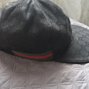 Svart Gucci keps  - Svart keps från Gucci med diskret GG-monogram över hela ytan. Klassisk böjd skärm och ikoniska gröna och röda ränder på sidan. Tillverkad i polyester och bomull, med etikett som visar 'Made in Italy'. Snygg och lyxig accessoar för dig som gillar designer.