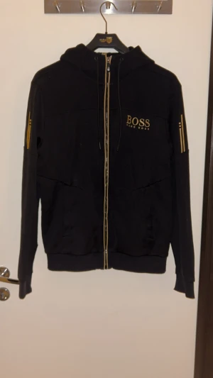 Svart hoodie från Hugo Boss med guldtryck - Svart hoodie från Hugo Boss med guldfärgade detaljer och logga på bröstet. Tröjan har huva med snörning, hel dragkedja framtill och ribbade muddar. Snygga guldtryck på ärmarna ger en exklusiv vibe. Perfekt för dig som gillar stilrena streetwear-plagg.