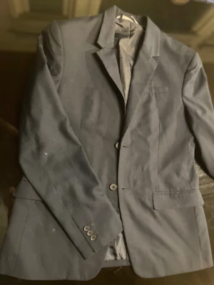 Mörkblå kostym med skjorta - Snygg mörkblå kostym med klassisk kavaj och matchande kostymbyxor. Kavajen har två knappar framtill, fickor med lock och stilren krage. Till kostymen ingår en mörkblå skjorta med knappar och liten broderad logga på bröstet. Perfekt för dig som vill ha en stilren look.