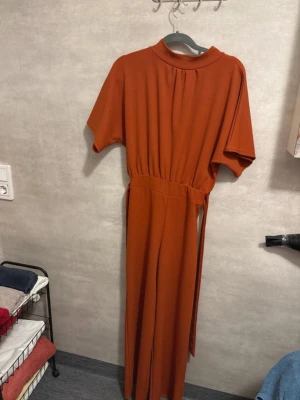 Rostfärgad byxdress med kort ärm - Säljer en snygg rostfärgad byxdress med korta ärmar och rundad hals. Byxdressen har ett brett midjeband med knyt i sidan och vida ben. Materialet känns mjukt och har ett fint fall, perfekt för dig som gillar bekväma och stilrena plagg.