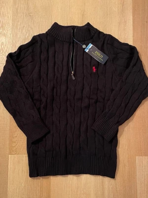 Svart stickad tröja Polo Ralph Lauren - Svart kabelstickad tröja från Polo Ralph Lauren med half zip och ribbad krage. Klassisk röd logobrodyr på bröstet. Långärmad och tillverkad i mjukt material som passar perfekt för kyliga dagar. Snygg och tidlös design med normal passform.