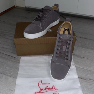 Grå vita sneakers från Christian Louboutin - Snygga grå sneakers från Christian Louboutin med vit platt sula och rund tå. Skorna har snörning och är tillverkade i mocka med beige innersula. Perfekt för dig som gillar stilrena och lyxiga detaljer. Kommer med originalkartong och dustbag.