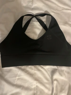 Svart sport-bh med korsade band - Svart sport-bh med korsade axelband och justerbar knäppning bak. Tillverkad i ett stretchigt syntetmaterial som ger bra stöd vid träning. Diskret logga framtill i svart. Perfekt för gymmet eller andra sportaktiviteter.