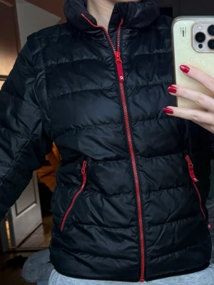 Svart pufferjacka med röda dragkedjor - Svart pufferjacka från Rukka med röda dragkedjor och hög krage. Jackan har quiltad design och två sidofickor med röda detaljer. Perfekt för dig som gillar sportig och clean stil. Passformen är normal och materialet känns lätt och smidigt.