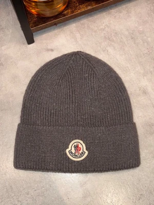 Grå Moncler mössa i ull - Säljer en snygg grå Moncler mössa i mjuk och ribbstickad ull. Mössan har en uppvikt kant och ett klassiskt Moncler-märke framtill. Perfekt för dig som vill ha en stilren och varm accessoar till vintern.