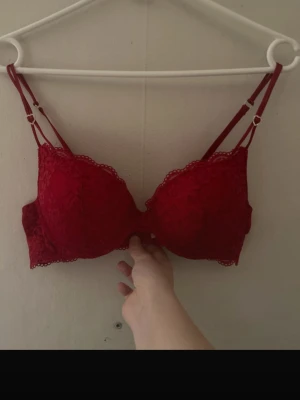 Röd spets-bh från Lindex  - Snygg röd bh från Lindex med spetsdetaljer och dubbla axelband. Axelbanden har små hjärtformade detaljer i metall och kuporna är vadderade för extra stöd. Perfekt för dig som gillar romantiska detaljer och vill ha något extra.