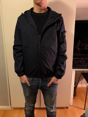 Marinblå vindjacka från Stone Island - Säljer en marinblå vindjacka från Stone Island med huva och dragkedja framtill. Jackan har den klassiska loggan på vänster ärm och två sidofickor med dragkedja. Perfekt för dig som gillar stilrena och sportiga plagg.