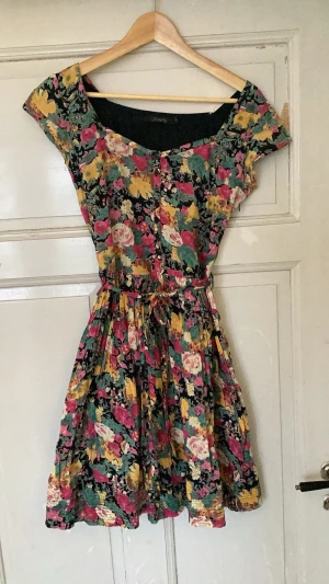 Blommig kortklänning med knyt - Säljer en färgglad kortklänning med blommigt mönster i rosa, gul, grön och svart. Klänningen har korta ärmar, rund hals och ett knytband i midjan som ger en snygg siluett. Perfekt för dig som gillar färg och mönster! Har en liten skada på urigningen i bak.