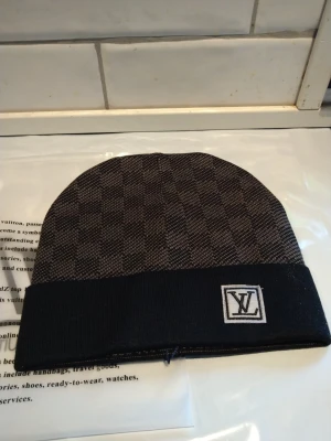 Rutig mössa från Louis Vuitton - Snygg mössa från Louis Vuitton med klassiskt rutigt mönster i mörkgrå och svart. Mössan har en bred svart kant med LV-logga framtill. Perfekt accessoar för att lyfta din stil under kyliga dagar.
