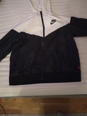 Nike vindjacka med huva, svart och vit - Snygg vindjacka från Nike i svart och vitt med huva och hel dragkedja framtill. Jackan har klassisk logga på bröstet och två sidofickor. Perfekt för dig som gillar sportig stil och vill ha något lätt att slänga på sig.