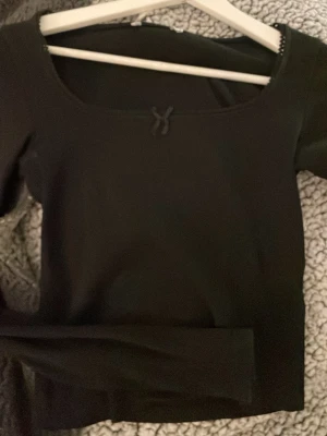 Svart långärmad topp med rosett - Säljer en svart långärmad topp med en liten söt rosett framtill vid halsringningen. Toppen har en bred, rundad urringning och är gjord i ett mjukt, ribbat material som sitter snyggt på kroppen. Perfekt att styla med jeans eller kjol.