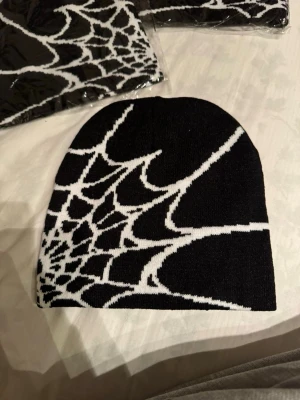 Svart och vit mössa med spindelnätsmönster - Cool svart mössa med vitt spindelnätsmönster som täcker hela ytan. Mössan är stickad och har en klassisk form som passar de flesta huvuden. Perfekt för dig som gillar edgy och unik stil. Snygg detalj som sticker ut i mängden.