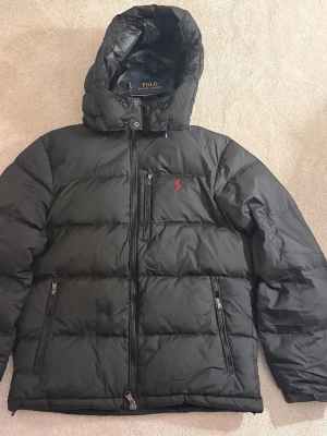 Svart pufferjacka från Polo Ralph Lauren - Säljer en svart pufferjacka från Polo Ralph Lauren med huva och dragkedja framtill. Jackan har två sidofickor med dragkedja och den klassiska röda Polo-loggan på bröstet. Perfekt för kalla dagar och riktigt snygg till streetwear.