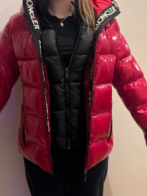 Röd pufferjacka med huva XS/S - Säljer en snygg röd pufferjacka med glansig finish och svart innerfoder. Jackan har huva med Moncler-logga, två stora dragkedjefickor och klassisk patch på ärmen. Perfekt för kalla dagar och ger en sportig vibe. Köpt från Plick men har inte använd, lite för liten. 