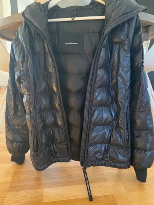 Svart Peak Performance dunjacka tomic strl S - Svart dunjacka från Peak Performance med huva och snygga quiltade paneler. Jackan har dragkedja framtill, praktiska fickor och en  logotyp på ärmen. Perfekt för kalla dagar och har en modern, lite loose passform.