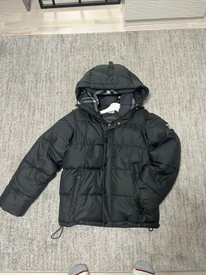 Svart pufferjacka från Burberry - Kommer med dustbag och blir till väst.   Säljer en svart pufferjacka från Burberry med huva och dragkedja. Jackan har Burberry-logga på ärmen och klassiskt rutigt foder i huvan. Perfekt för kalla dagar och riktigt snygg streetstil. Två fickor med dragkedja och justerbar nederkant.