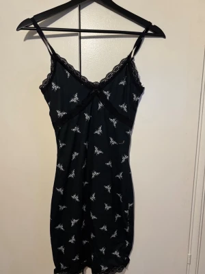 Svart kort klänning med spets - Svart kort klänning med tunna axelband och spetsdetaljer vid halsringning och nederkant. Klänningen har ett vitt fjärilsmönster över hela tyget och är figurnära med omlottliknande skärning vid bysten. 
