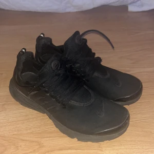 Svarta Nike Presto sneakers - Säljer ett par helsvarta Nike Presto sneakers med klassisk mesh-överdel och plastdetaljer på sidorna. Skorna har snörning, rund tå och platt sula för skön komfort. Perfekta för dig som gillar stilrena och sportiga skor med en clean look.