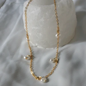 Guldfärgat halsband och armband med pärlor - Säljer ett halsband i guldfärgad kedja dekorerade med vita pärlor. Kedjan är tunn och elegant, och pärlorna är oregelbundna för en modern look. Perfekt accessoar för att lyfta din outfit. Priset för både halsbandet och armbandet är 100kr. 
