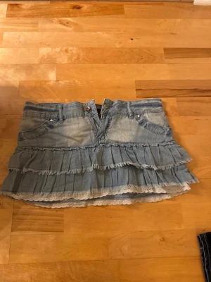 Kort lågmidjade volangkjol i jeans - Säljer en kort lågmidjad jeanskjol i ljusblå denim med flera volanger och spetsdetalj längst ner. Perfekt för dig som gillar en söt och cool stil. Midjemåttet är 39 cm och längden 27 cm💞pris går att diskutera! 