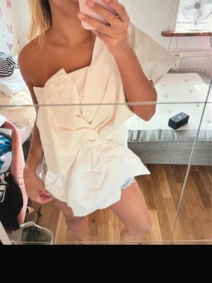 Vit topp med rosett - Säljer en vit oneshoulder topp med rosett framtill. Toppen har asymmetrisk skärning och är axelbandslös på ena sidan. Knappt använd o är i storlek xs men skulle säga mellan xs/s