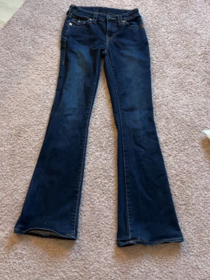 True Religion blå bootcut jeans - Snygga mörkblå jeans från True Religion med bootcut passform och klassiska fickor med guldfärgade knappar och kontrastsömmar. Jeansen har detaljerad brodering på bakfickorna och är tillverkade i jeansmaterial med midwaist. Perfekt för dig som gillar ikoniska streetwear-vibbar.