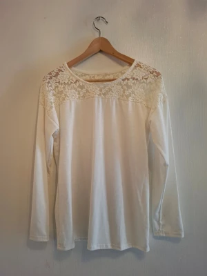 Vit långärmad blus med spets  - Säljer en vit långärmad blus med vackra spetsdetaljer och blommigt mönster över axlar och bröst. Blusen har rund halsringning och är tillverkad i mjukt material som känns skönt mot huden. Perfekt för dig som gillar romantiska detaljer och stilren look.