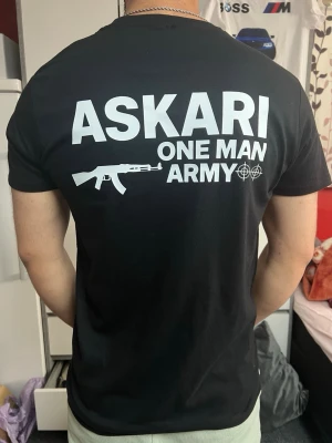 Svart ASKARI t-shirt med tryck - Svart t-shirt från ASKARI med vit text och grafik både fram och bak. Trycket visar 'ASKARI ONE MAN ARMY' tillsammans med en gevärsikon och sikte. T-shirten har rund hals och korta ärmar, och är gjord i mjuk bomull som sitter skönt mot kroppen.