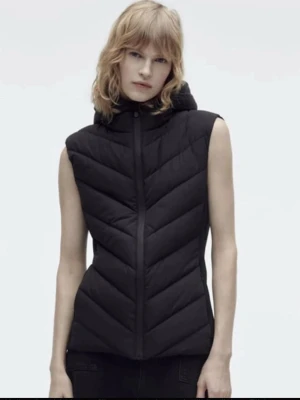 Svart dunväst med huva - Snygg svart dunväst med huva och dragkedja framtill. Västen har ett quiltat mönster i sicksack och är ärmlös, perfekt att slänga över en hoodie eller tröja. Enkel och stilren design som passar till många outfits.