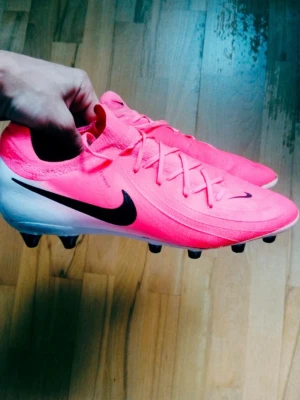 Nike Phantom Gx Ag Pro - Säljer ett par Nike Phantom fotbollsskor i en riktigt snygg rosa färg med vita och svarta detaljer. Skorna har snörning, rund tå och platt sula med dobbar för gräsplan. Ovandelen är i syntetmaterial och designen är modern och sportig. Perfekt för dig som vill sticka ut på planen. Skor för konstgräs 