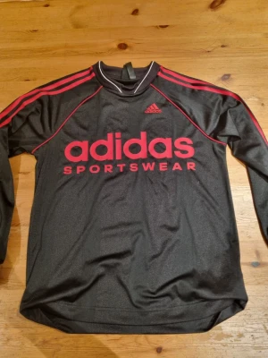 Svart långärmad Adidas tröja - Svart långärmad tröja från Adidas med röda detaljer och tryck på bröstet. Klassisk sportig look med röda ränder längs axlarna och ribbad krage med vit kant. Perfekt för träning eller chill. Materialet är troligtvis polyester och har en mjuk känsla.