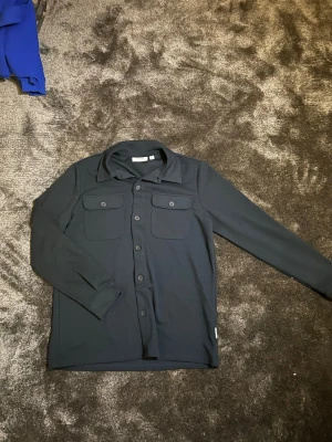 Mörkblå overshirt från Only & Sons - Snygg mörkblå overshirt från Only & Sons med klassisk krage och två bröstfickor med knappar. Jackan har långa ärmar och knäppning hela vägen framtill. Perfekt att slänga på över en t-shirt för en chill look.