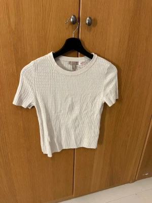 Ribbad vit topp från H&M - Säljer en ribbad vit topp från H&M i storlek M. Toppen har korta ärmar, rund halsringning och ett diskret hålmönster i tyget. 