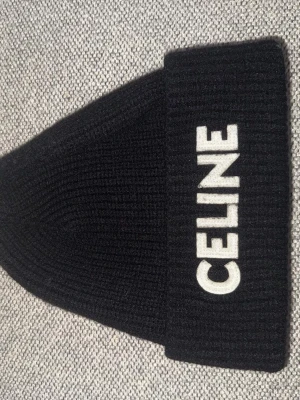 Celine mössa - Snygg svart ribbstickad mössa från Celine med vit broderad logga framtill. Mössan har en bred uppvikt kant och är gjord av ull som håller dig varm. Perfekt för dig som vill ha en clean och lyxig look.