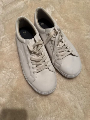 Vita sneakers  - Säljer ett par stilrena vita sneakers från H&M i storlek 43. Skorna har klassisk rund tå, platt sula och snörning framtill. Tillverkade i syntetmaterial med diskreta sömmar och perforerade detaljer på sidorna. Perfekta för dig som gillar enkel och clean stil.