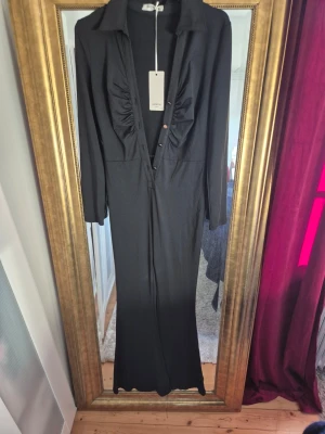 Svart byxdress från Giorgia Paris - Svart långärmad byxdress med djup v-ringning och knappar framtill. Byxdressen har krage och rynkade detaljer vid bysten. Lång, rak passform och tillverkad i ett mjukt, stretchigt material.