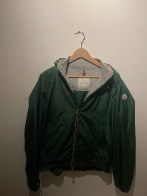  (Äkta) Grön vindjacka från Moncler -  Säljer en moncler som är 100 procent äkta.  Men den är lite mindre i storlek för den är krympt i tvätten så passar för dig som är 165 till 176. Snygg grön vindjacka från Moncler med huva och coola detaljer som röd-vit rand vid dragkedjan. Jackan har långärmade ärmar, två fickor och Moncler-logga på ärmen. Insidan av huvan är grå och materialet känns lätt och smidigt. Perfekt för dig som gillar streetstyle.
