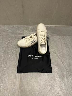 Saint Laurent vita sneakers med broderi - Snygga vita sneakers från Saint Laurent med handskriven broderad logga på sidan. Skorna har rund tå, platt sula och snörning. Tillverkade i canvas med detaljer i skinn och levereras med svart dustbag med Saint Laurent Paris-tryck. Storlek 41 och dustbag medföljer!