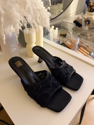 Svarta sandaler med låg klack, strl 38 - Säljer ett par svarta sandaler med låg klack och öppen tå. Ovandelen har ett snyggt blommigt spetsmönster och sandalerna har en fyrkantig tå. Perfekta för dig som gillar stilrena och trendiga skor med lite extra detaljer.