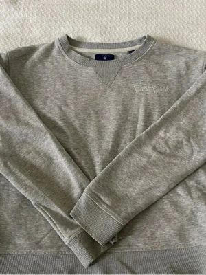 Grå sweatshirt från Gant - Klassisk grå sweatshirt från Gant med broderat på bröstet. I mycket gott skick. Tröjan är i storlek 170/15 år, skulle säga storlek S.
