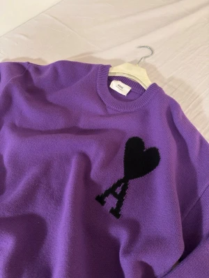 Lila stickad tröja från Ami - Säljer en lila stickad tröja från Ami Alexandre Mattiussi med svart hjärta och bokstaven A på bröstet. Tröjan har rund hals, ribbade muddar och är långärmad. Materialet är mjuk ullblandning som känns riktigt skön mot huden.