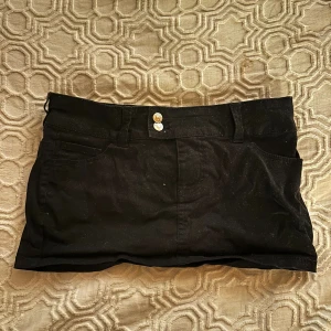 Svart kortkjol med fickor - Säljer en svart kortkjol med klassisk jeanslook, utrustad med både fram- och bakfickor samt bälteshällor. Kjolen har två knappar framtill och är gjord i ett stadigt bomullsmaterial. Perfekt för dig som gillar enkel och stilren design.