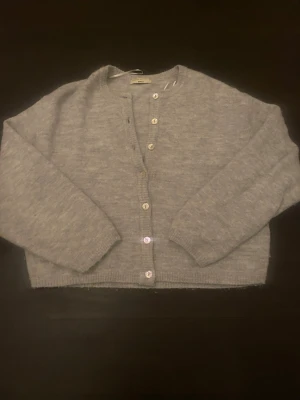 Grå stickad kofta från gina - Mysig grå stickad kofta från gina med rund hals och knappar framtill. Koftan har långärmade ärmar och ribbade muddar vid ärmslut och nederkant. Perfekt att slänga på sig när det är lite kyligt ute.