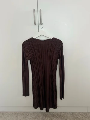 Plisserad mörkbrun klänning Boohoo - Säljer en snygg plisserad mörkbrun kortklänning från Boohoo. Klänningen har lång ärm, rund halsringning och är gjord i ett glansigt, lätt material som ger en cool struktur. Perfekt för dig som vill sticka ut med en trendig look!