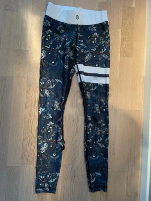 Mönstrades leggings från Stronger S - Supersnygga leggings från Stronger i storlek S med mörkblå botten och ugglemönster samt vita detaljer i midjan och på benet. Tillverkade i stretchigt material med 82% polyester och 18% elastan. Perfekta för träning eller chill.