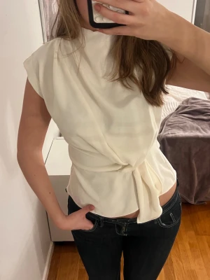 Vit omlottopp med knyt från Zara - Säljer en stilren vit omlottopp från Zara i storlek XS. Toppen har hög hals, är ärmlös och har en snygg knytning i midjan som ger en feminin siluett. Materialet känns mjukt och lätt. Passar Xs - M